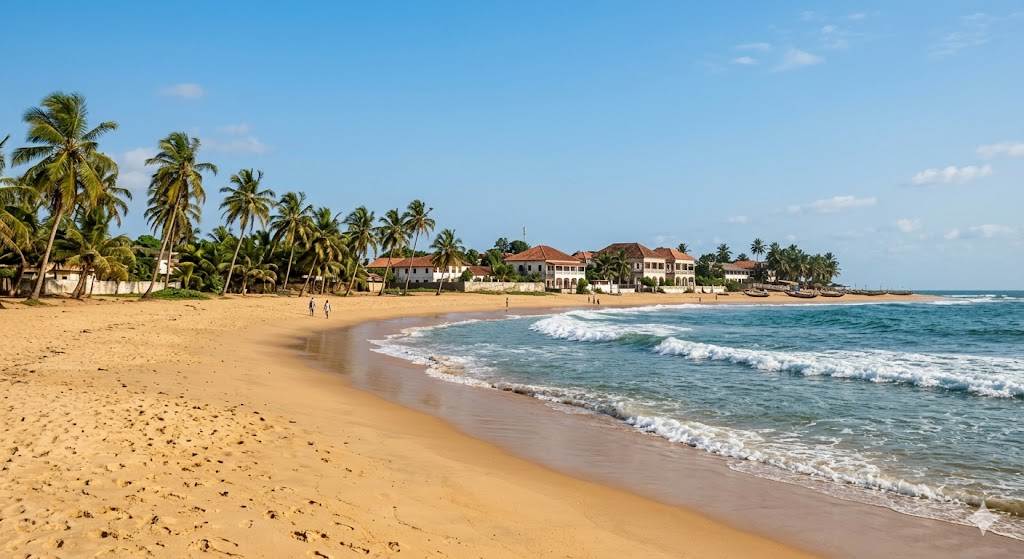 A beautiful landscape photograph of Grand-Bassam beach during the dry season in January. Gentle ocean waves roll onto clean golden sand, palm trees sway under a clear blue sky, and colonial-era buildings are visible in the background under perfect weather conditions.